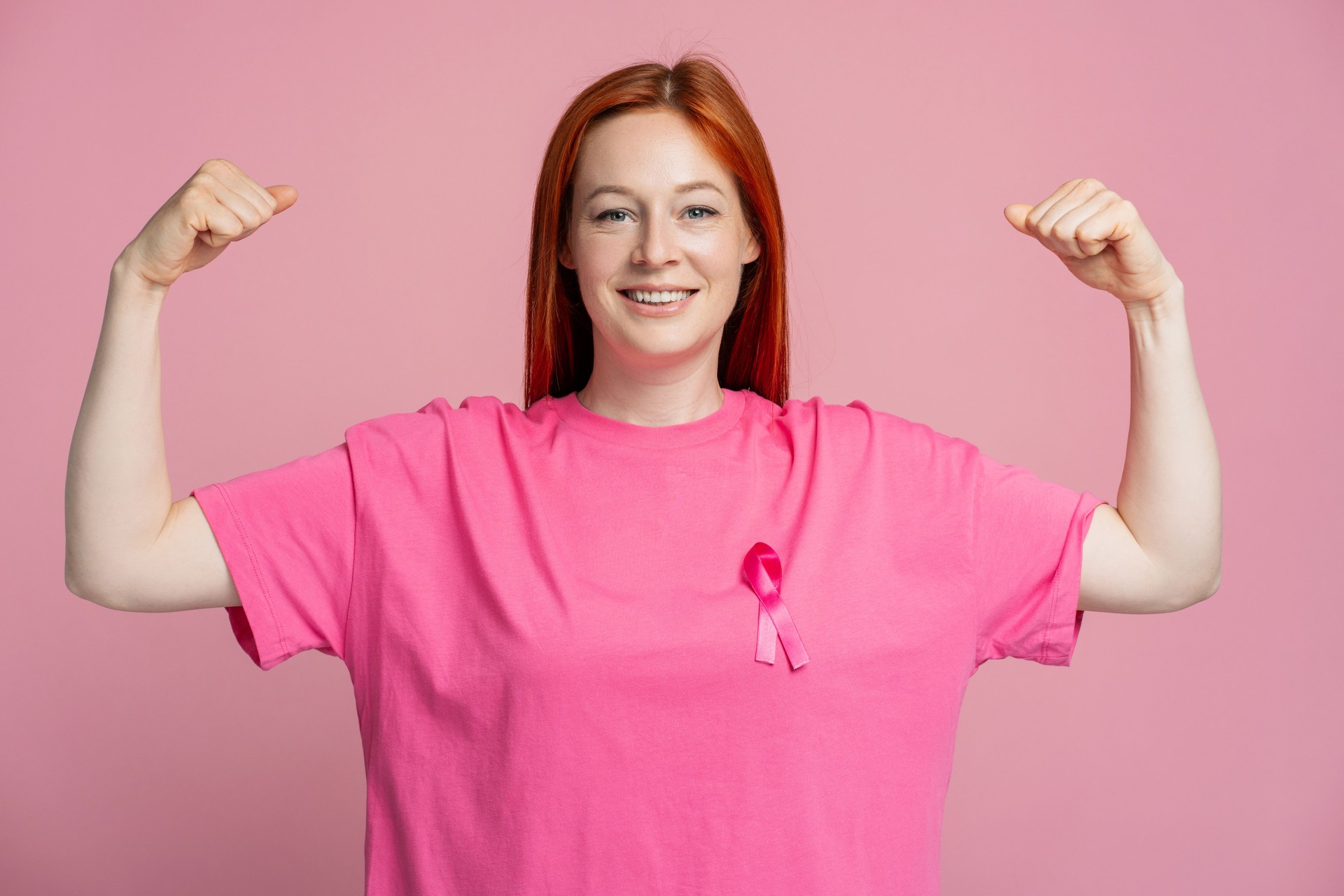 Smiling woman wearing pink t shirt with pink ribbon showing biceps isolated on pink background