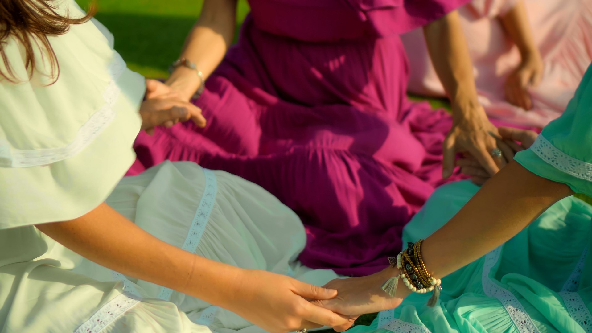 European girls friends in dresses sitting in a circle holding each other's hands in nature with green grass on the background